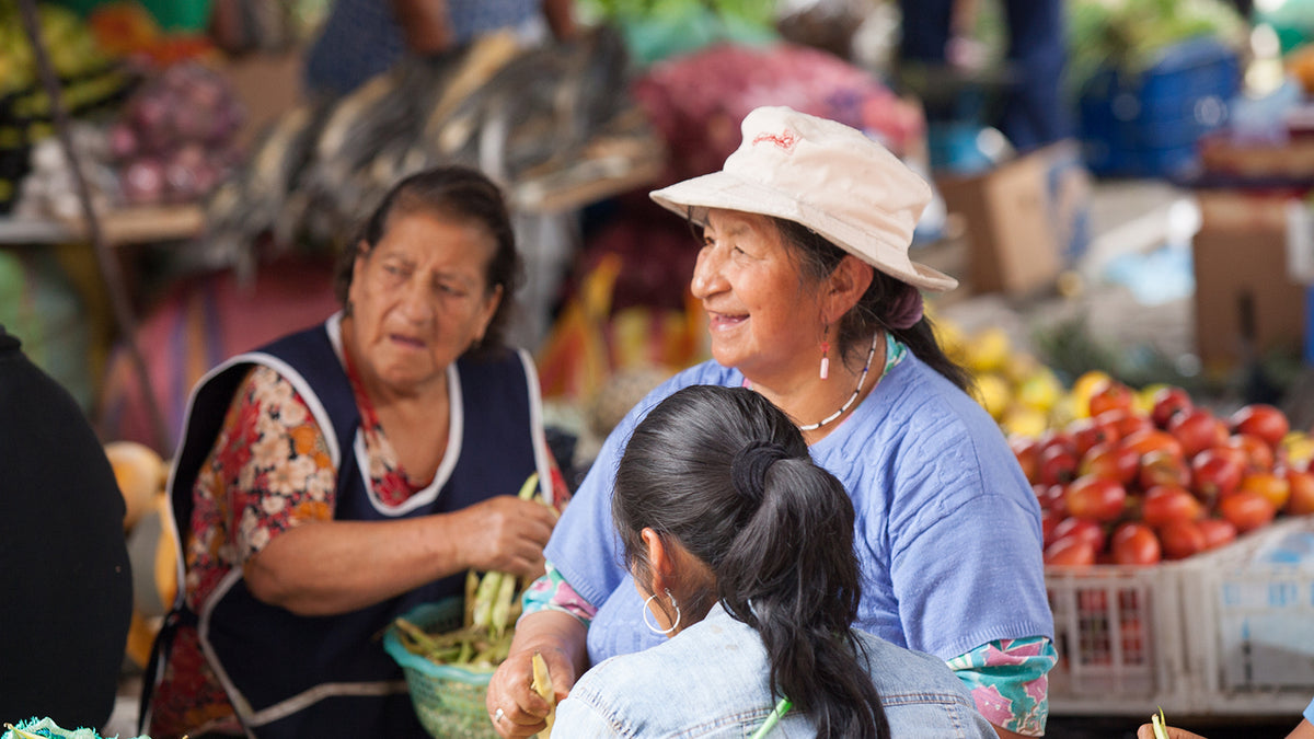 Image of an ecuadorian woman wearing a blue t-shirt and a white sunhat. She is smiling and is sat with other women in a market.